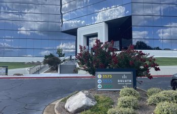 Office building with reflective glass facade, surrounded by landscaping. A sign in the foreground displays multiple addresses: 3575, 3555, and 3505 Duluth.
