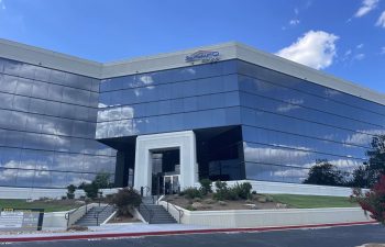 Modern office building with reflective glass facade under a blue sky with clouds. Stairs and landscaped greenery at the entrance.