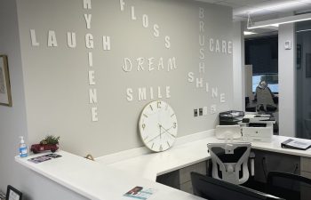 Dental office reception area with words like "Floss," "Care," and "Smile" on the wall. A clock, desk with a phone, computer, and a hand sanitizer bottle are visible.
