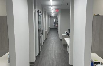 A long, narrow hallway with white walls and gray flooring, featuring overhead fluorescent lights and office spaces on each side.