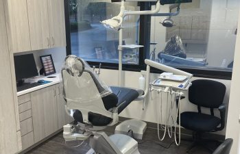 A modern dental office with an empty dentist's chair, dental equipment, and a monitor. Large window overlooking trees.