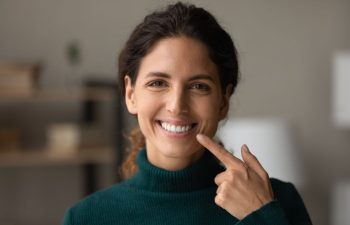 Woman with dark hair in a green turtleneck sweater smiling and pointing to her teeth, standing indoors with a blurred background.