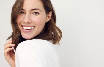 A smiling woman with brown, curly hair wearing a white top, with her hand touching her hair, against a light gray background.
