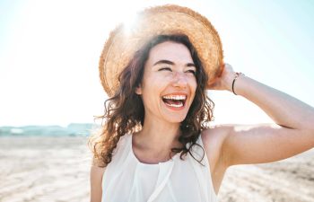 A woman with curly hair smiles broadly while holding a straw hat on her head, standing outdoors on a sunny beach.