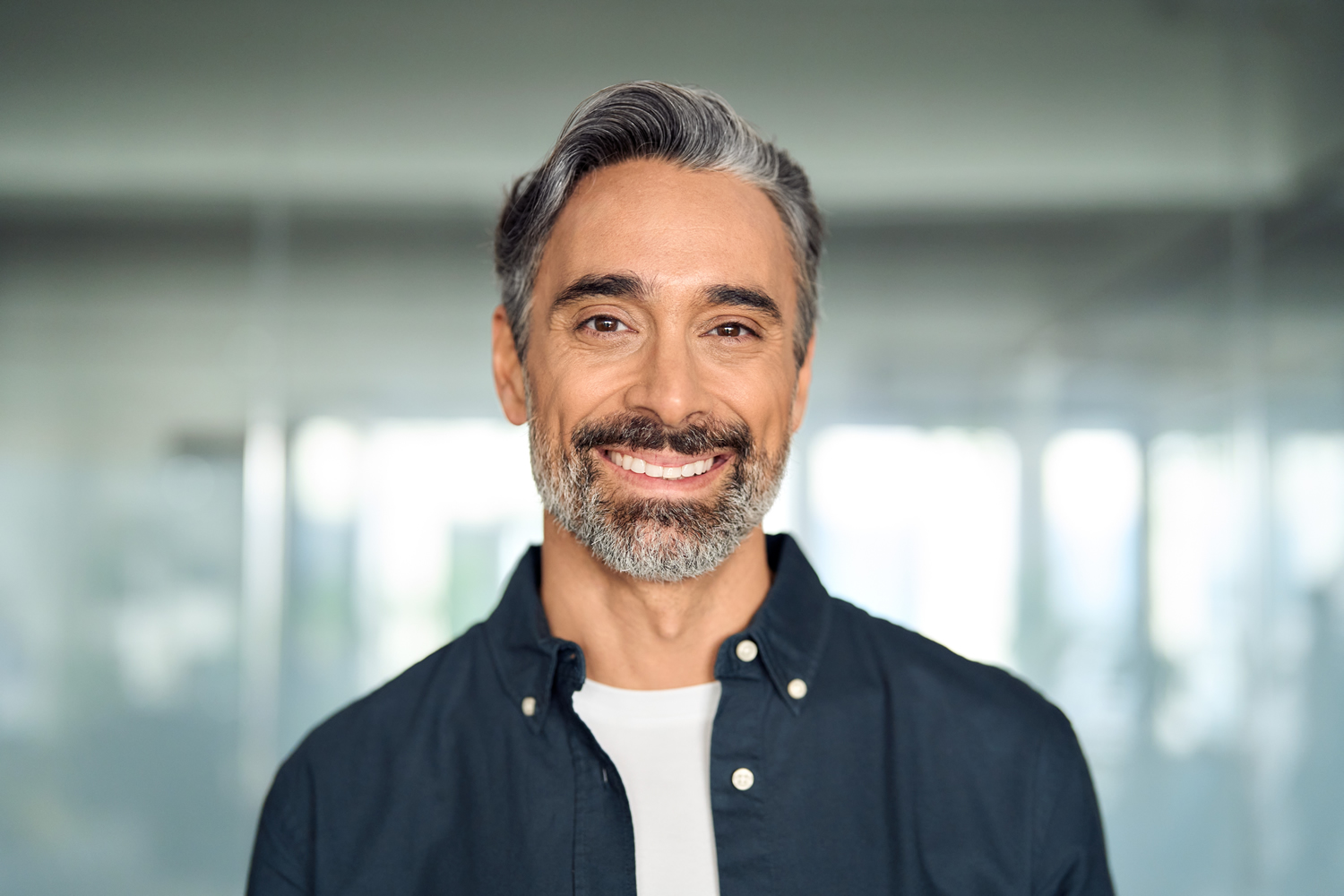 A headshot of a handsome, middle-aged man with salt-and-pepper hair and a beard, smiling warmly at the camera. He is wearing a dark blue button-down shirt over a white t-shirt, with a blurred modern office environment in the background.