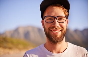 A bearded man wearing glasses and a cap smiles outdoors with mountains and a blue sky in the background.