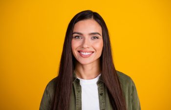 A smiling woman with long, straight dark hair and light-colored eyes, wearing a white t-shirt and an olive-green overshirt, looks at the camera against a solid yellow background.