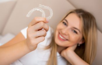 A close-up shot focusing on a woman's hand holding a clear dental aligner, with her smiling face blurred in the background.