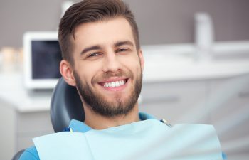 Portrait of a smiling young man with a beard sitting in a dental chair. He is wearing a blue bib and looking directly at the camera with a happy expression